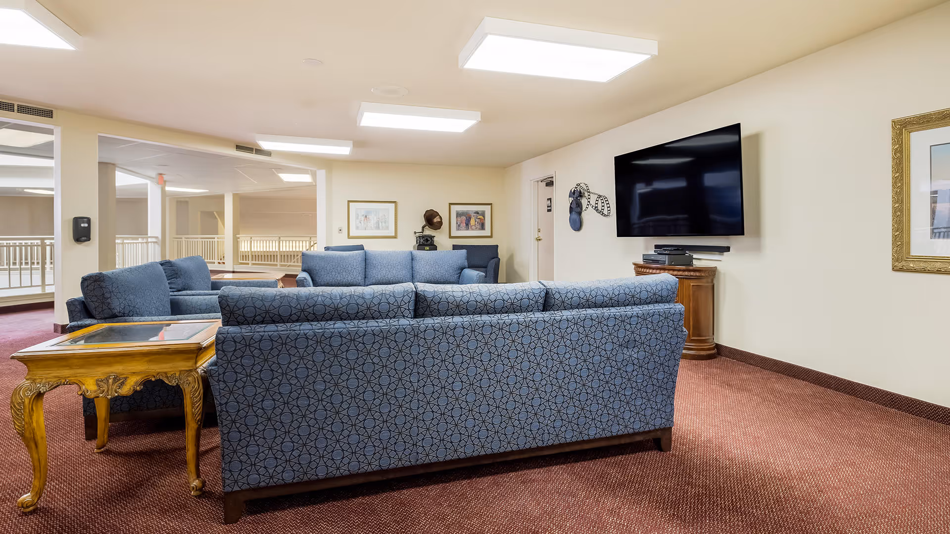 A cozy living room area with blue patterned sofas arranged around a wooden side table. A large flat-screen TV is mounted on the wall above a wooden stand. The room has beige walls, framed artwork, and a carpeted floor in a reddish-brown color. Ceiling lights illuminate the space, and there is an open area with railings visible in the background.