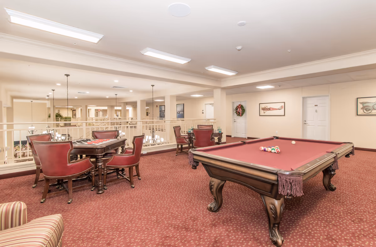 Bright carpeted game room in a senior living facility with a red-felt pool table, card table and red leather chairs.