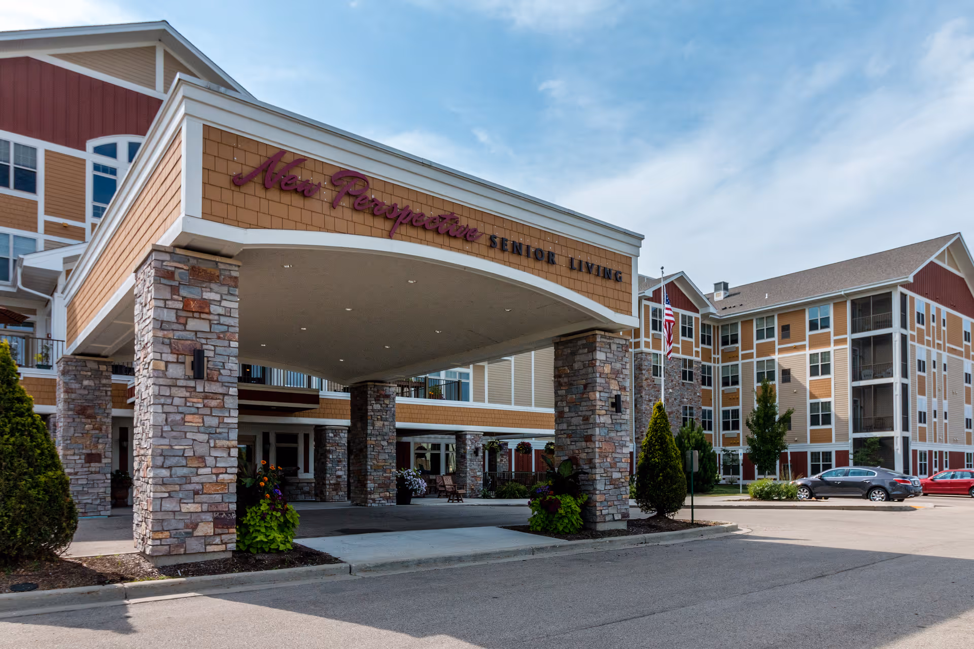Exterior view of New Perspective Senior Living facility showing the entrance with stone pillars and a covered driveway, surrounded by a parking area and multi-story residential buildings under a partly cloudy sky.