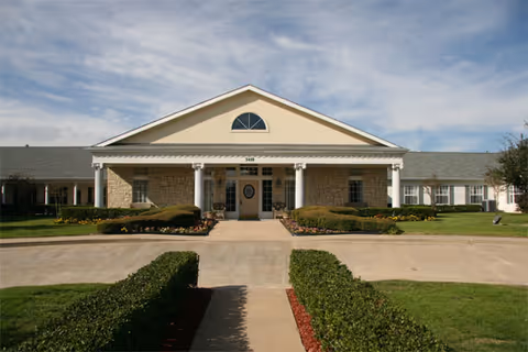 Front entrance of a single-story building with a columned portico, circular window in the door, and manicured lawns and hedges.