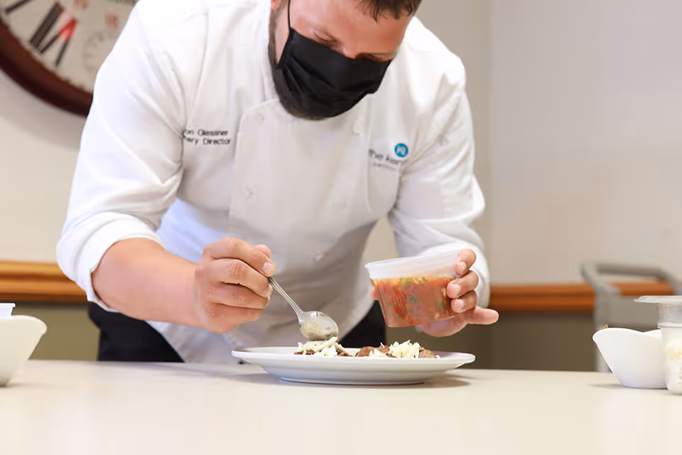 A chef wearing a white coat and black face mask is carefully spooning sauce from a plastic container onto a plated dish in a kitchen or food preparation area.