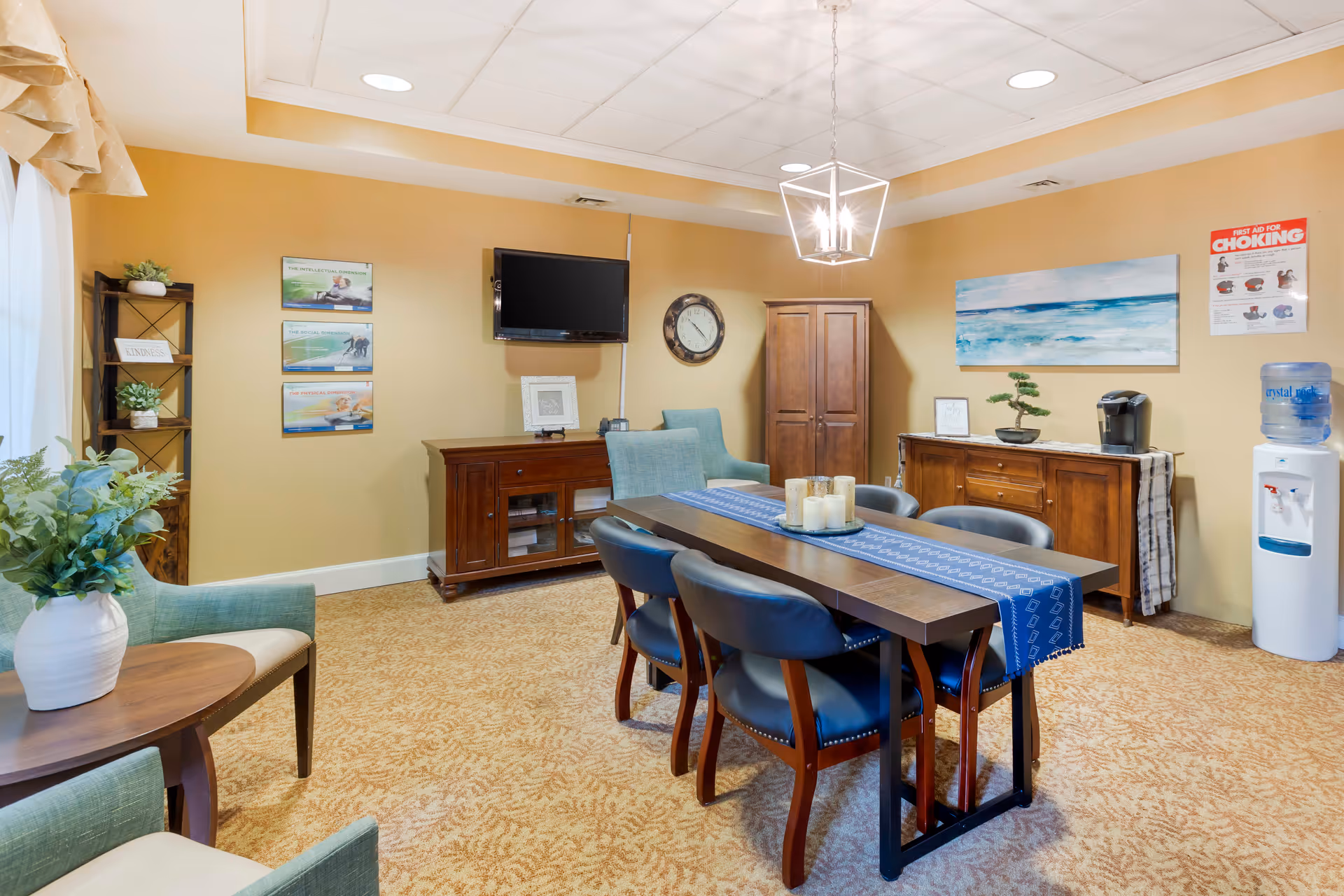 A cozy common area with a rectangular wooden table covered by a blue runner and surrounded by six blue cushioned chairs. The room has beige walls and carpet, a wall-mounted TV, a clock, wooden cabinets, a water dispenser, and decorative plants. There are also framed pictures and a first aid for choking poster on the walls.