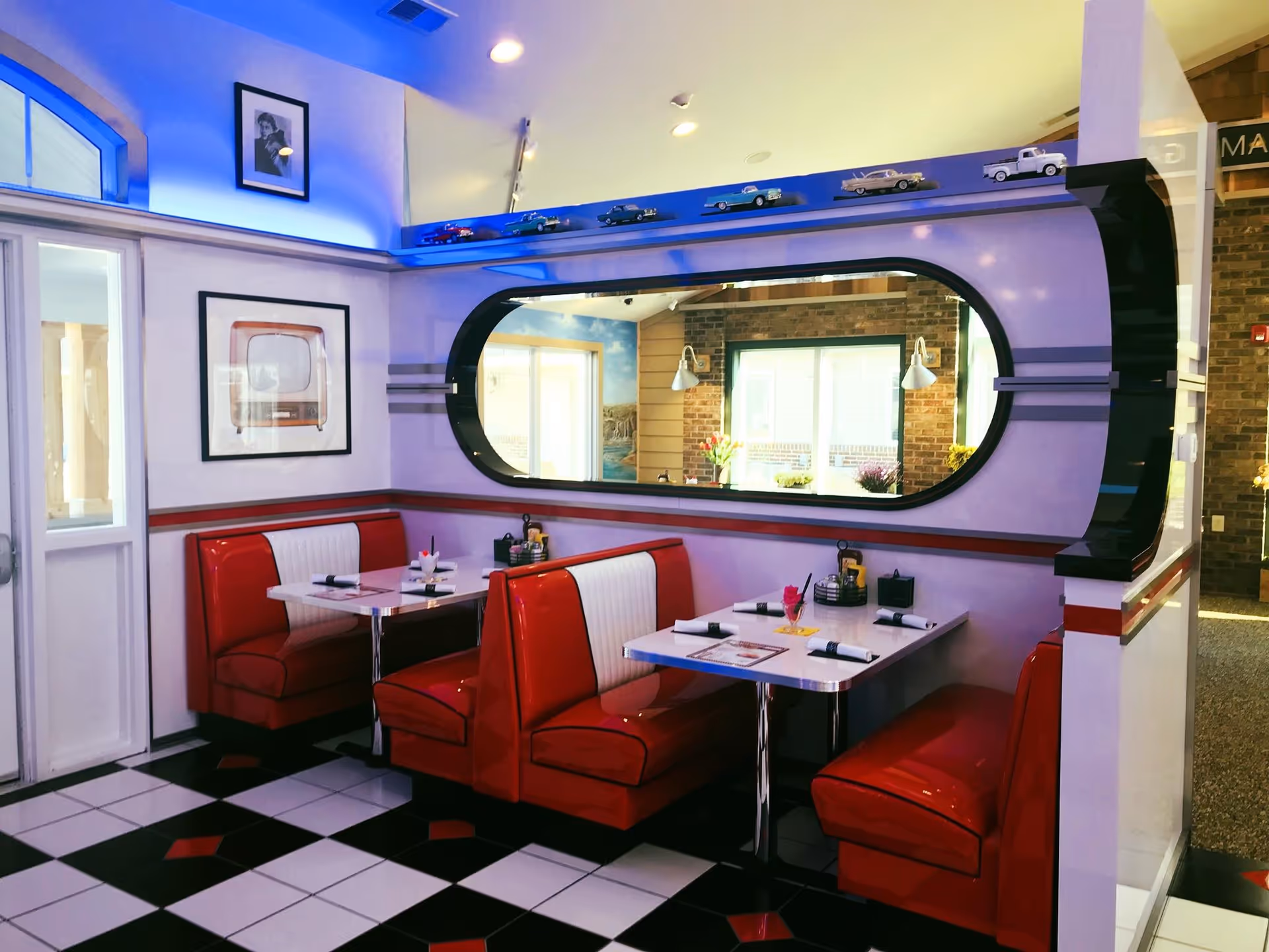 Retro diner-style communal dining area with red and white booths, checkered floor, and an oval mirror/window set into the wall.