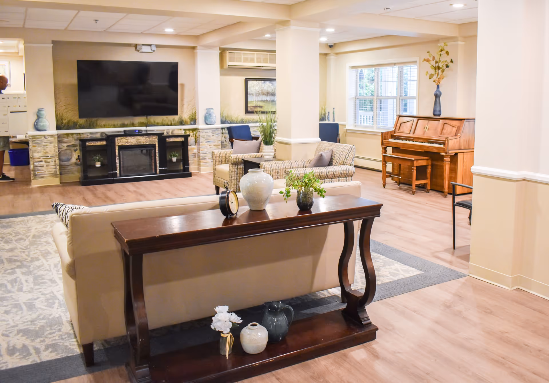 A bright and spacious senior living common area with a beige sofa, a wooden console table with decorative vases and a clock, two patterned armchairs, a large flat-screen TV mounted above a black fireplace, a wooden piano with a vase on top, and large windows letting in natural light.
