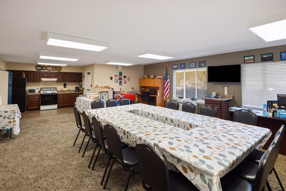A large room with multiple tables covered in patterned tablecloths and surrounded by black chairs. The room features a kitchenette area with dark wood cabinets, a stove, microwave, and refrigerator. There is an American flag near a window with blinds, a wall-mounted TV, and several framed pictures above the windows. The room has carpeted flooring and bright overhead lighting.