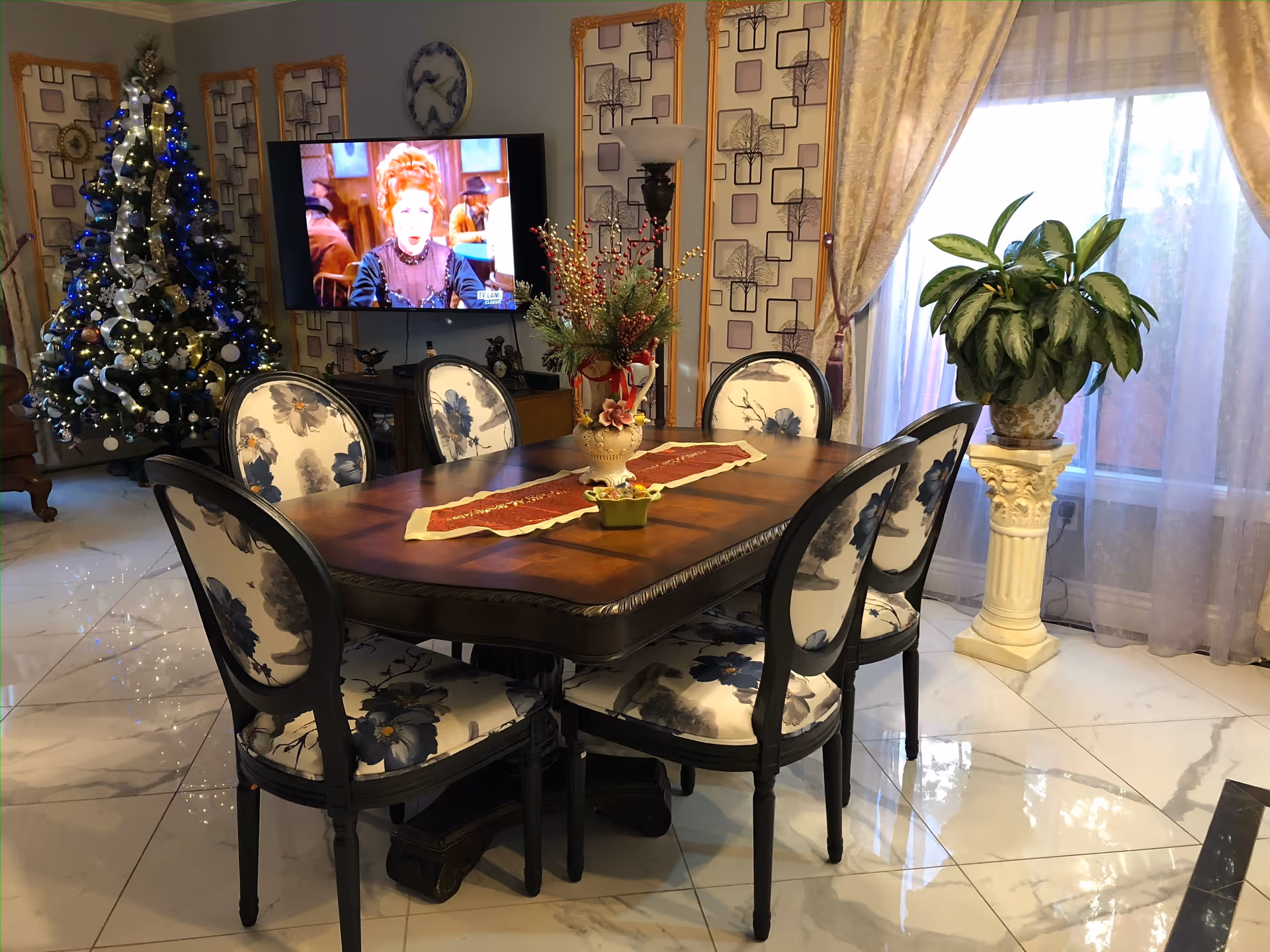 A dining room with a wooden table surrounded by six chairs with floral upholstery. The table has a red and white table runner and a decorative flower arrangement in the center. In the background, there is a decorated Christmas tree with blue and white lights, a wall-mounted TV showing a woman in period costume, patterned wallpaper, a tall floor lamp, and a large window with sheer curtains and a potted plant on a pedestal.