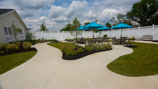 Sunlit courtyard with winding concrete paths, patio tables under turquoise umbrellas, landscaping, and a white privacy fence.