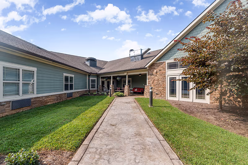 Concrete walkway leading to the covered entrance of a single-story building with stone siding, windows, seating, and lawns under a blue sky.