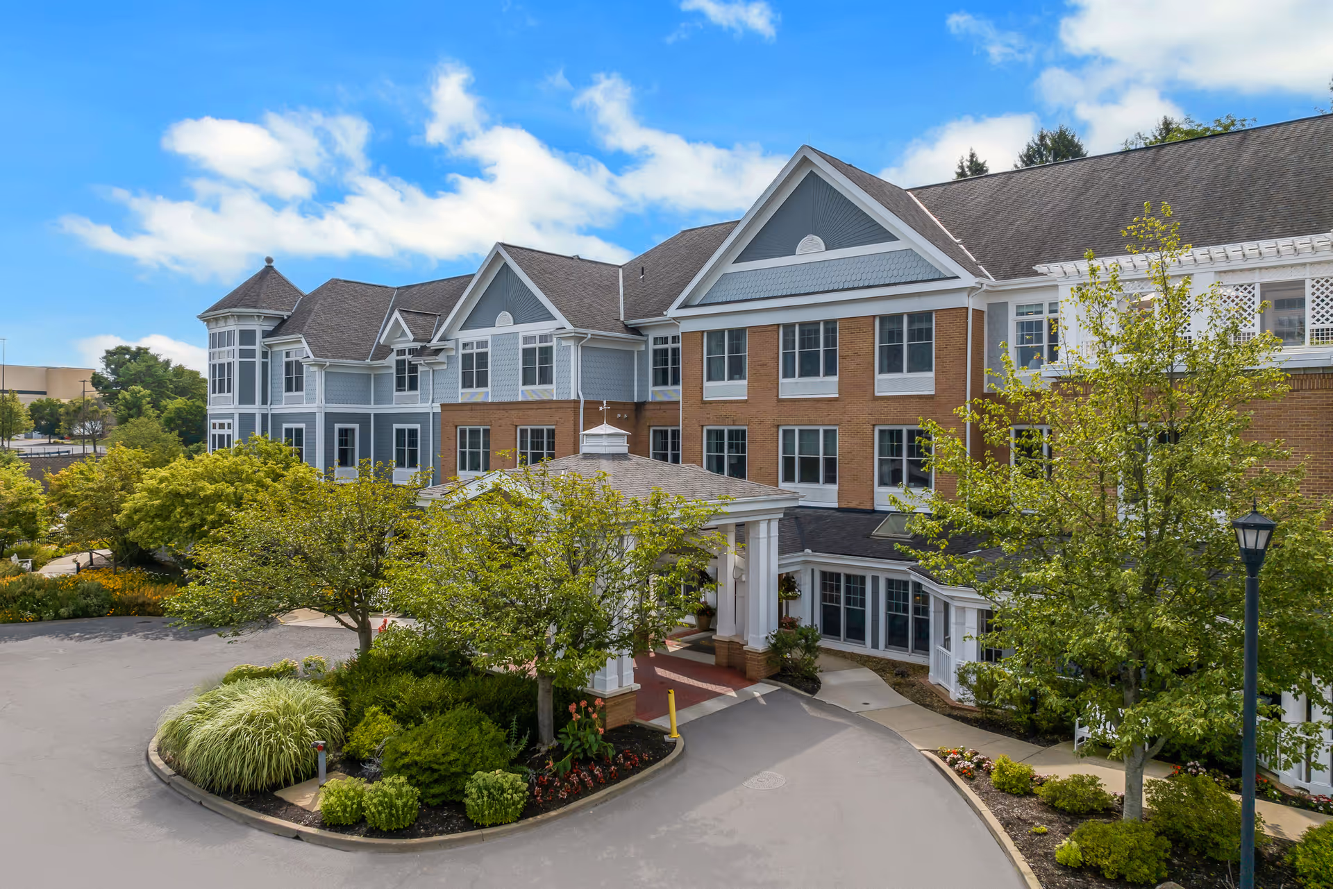 Three-story senior living building with a covered entrance, landscaped trees and a driveway under a blue sky.