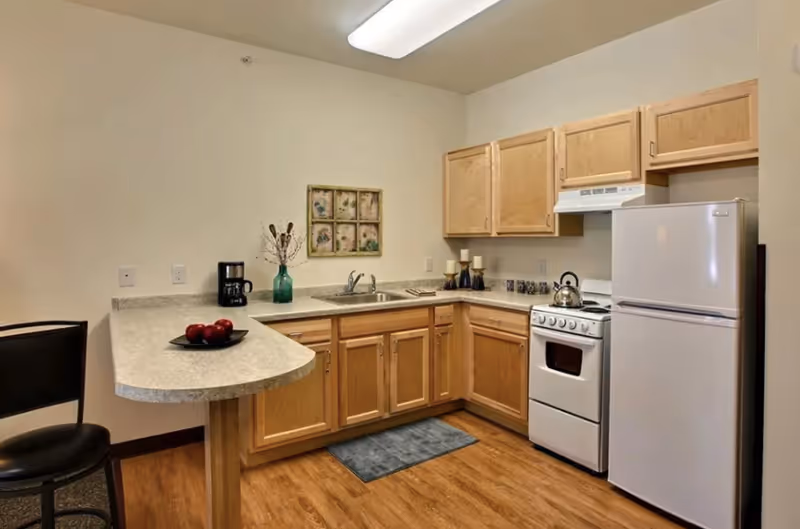 A small kitchen area with light wood cabinets, a white refrigerator, a white stove with an oven, a countertop with a sink, a coffee maker, a decorative vase with dried plants, and a plate with red apples. There is a black chair at the counter and a framed floral artwork on the wall.