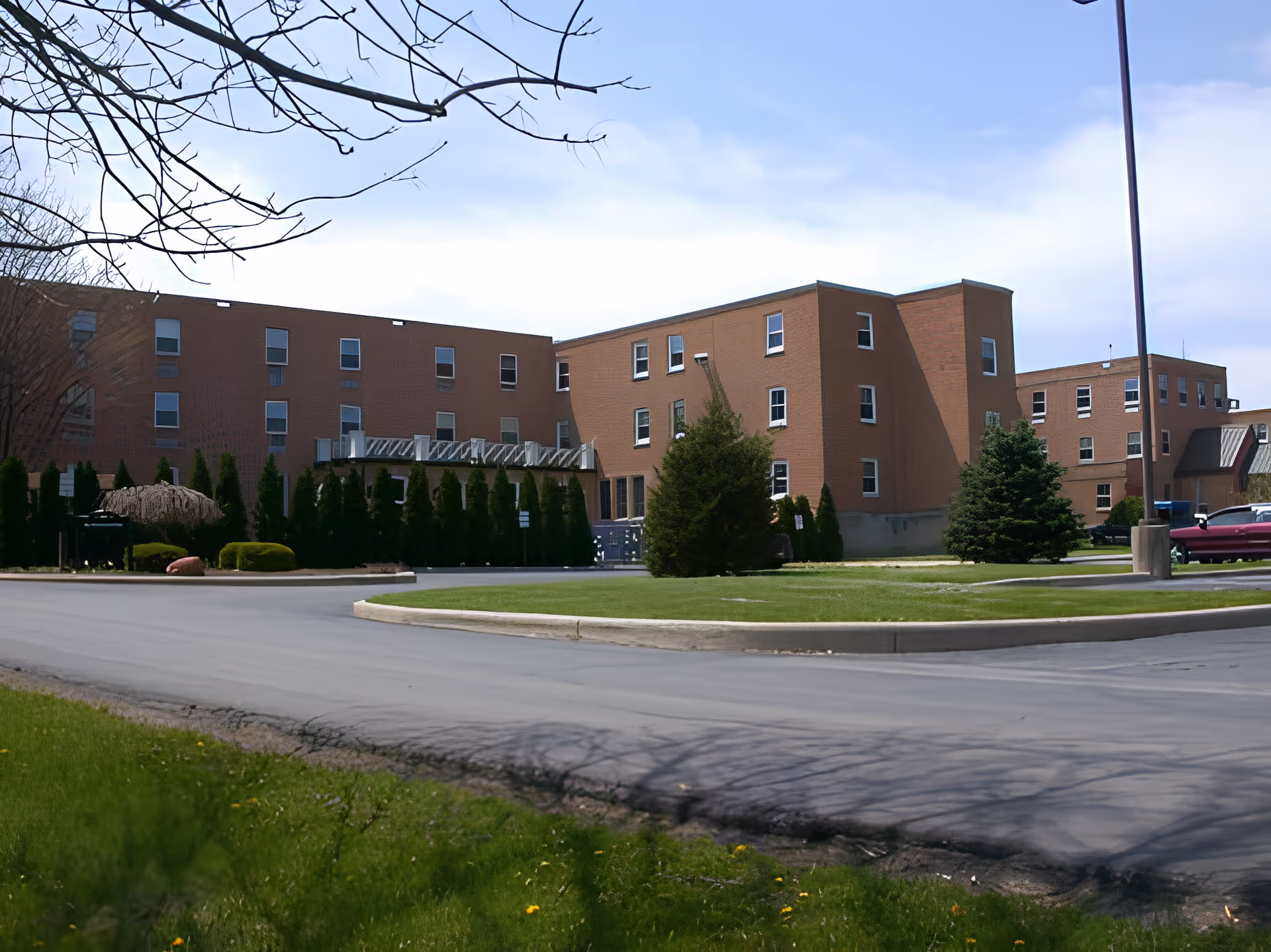 Exterior view of a multi-story brick building with several windows, surrounded by trees and shrubs, with a paved driveway and grassy areas in the foreground under a partly cloudy sky.
