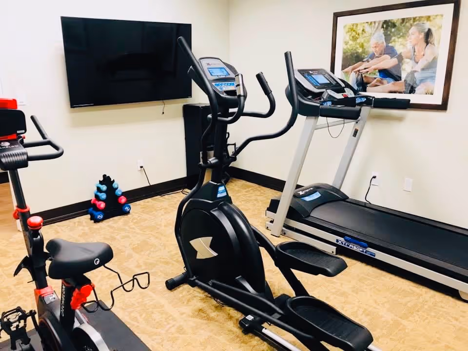 A small fitness room with exercise equipment including a stationary bike, an elliptical machine, and a treadmill. There is a wall-mounted flat screen TV and a rack of colorful dumbbells in the corner. A framed picture of two people exercising outdoors is hung on the wall.