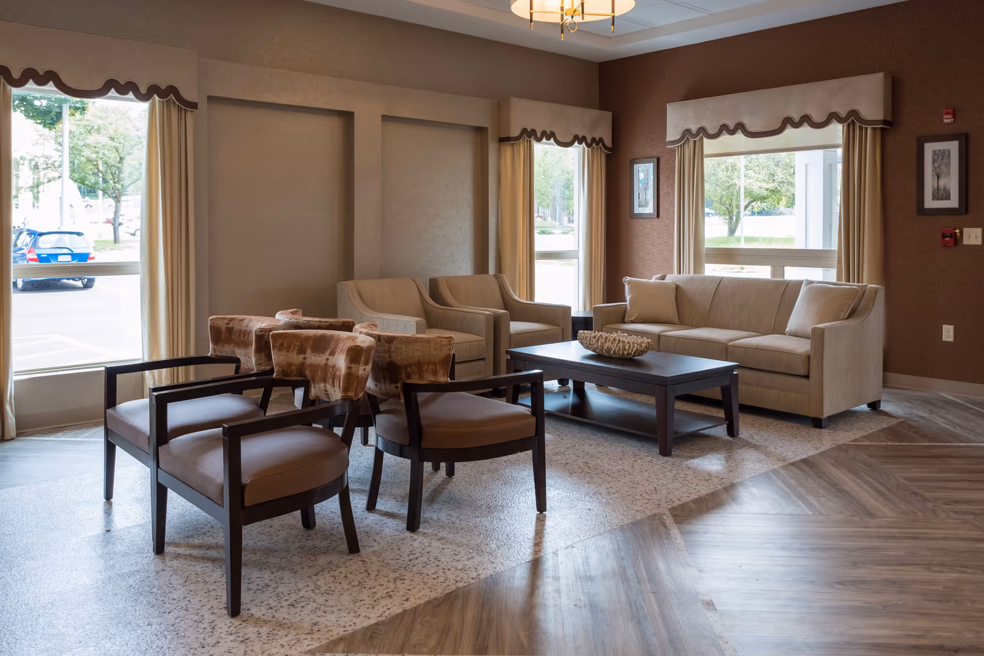 A cozy living room area with a beige sofa, two beige armchairs, and four brown cushioned chairs arranged around a dark wooden coffee table. The room has large windows with cream-colored curtains and valances, allowing natural light to brighten the space. The floor features a combination of speckled and wood-patterned flooring, and the walls are painted in warm tones with framed artwork.