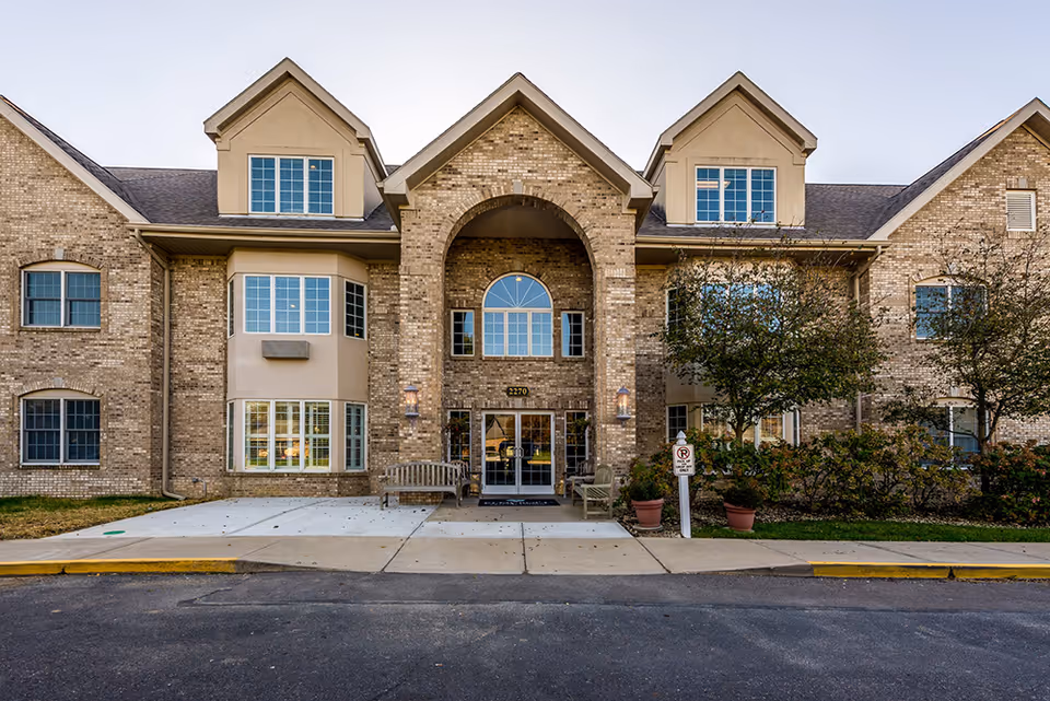 Front exterior view of a two-story assisted living facility building with brick and beige siding, multiple windows, an arched entrance with glass doors, benches on either side of the entrance, and some landscaping including trees and potted plants.