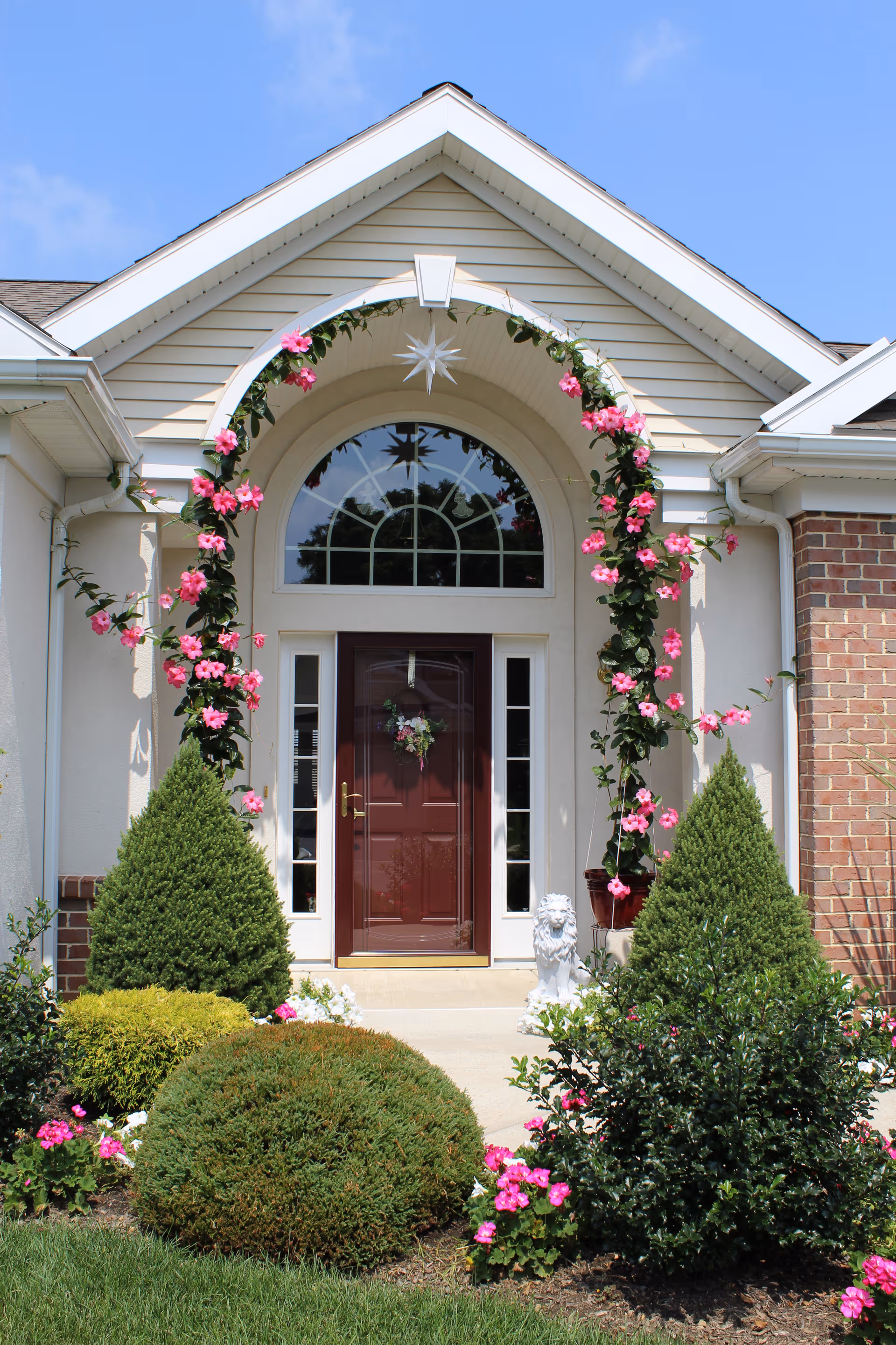 Front entrance of a building with a maroon door framed by white sidelights and a large arched window above. The entrance is decorated with a floral arch of pink flowers and greenery. There are neatly trimmed bushes and flowering plants in the garden bed leading up to the door, and a small white lion statue is positioned near the entrance. The sky is clear and blue.