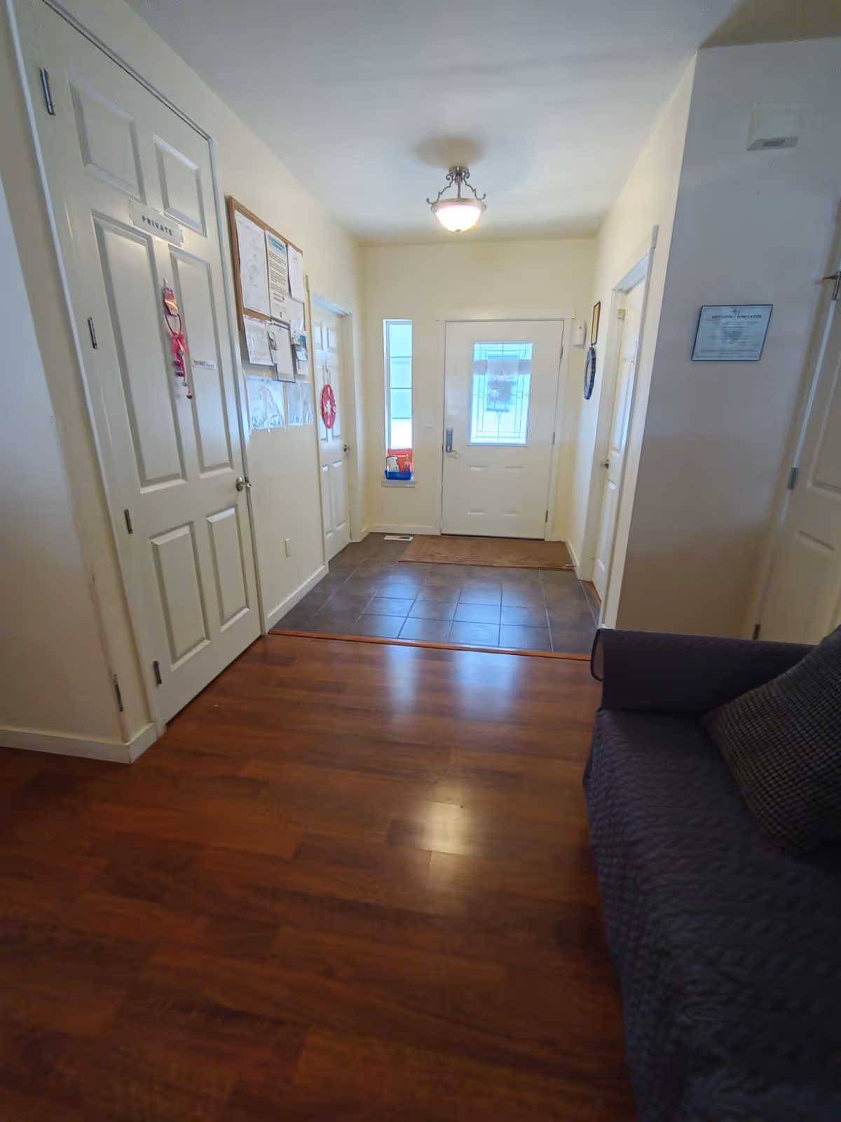 Interior view of a hallway in a senior living facility with wooden flooring leading to a tiled entryway with a white front door and side window. There are multiple white doors along the hallway, a bulletin board with papers on the left wall, and a dark-colored couch with a cushion on the right side.