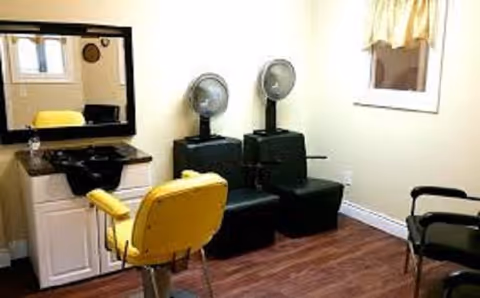 Interior salon area with a yellow barber chair, a sink and mirror, and two hooded hair dryers on dark chairs.