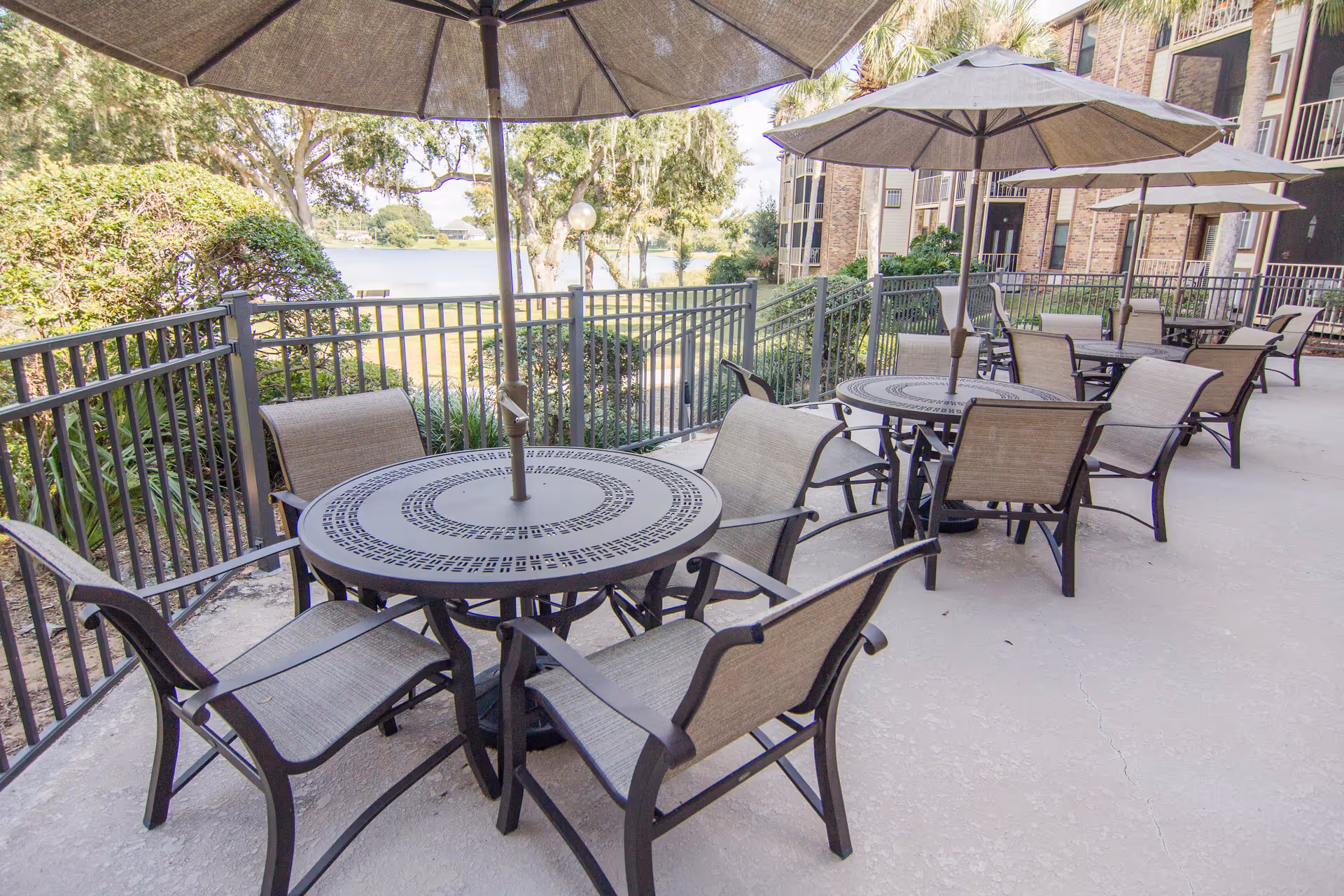 Outdoor patio area with multiple round metal tables and beige mesh chairs arranged around them. Each table has a large beige umbrella providing shade. The patio is enclosed by a black metal fence, and there are trees, bushes, and a lake visible in the background. A multi-story brick building is adjacent to the patio.