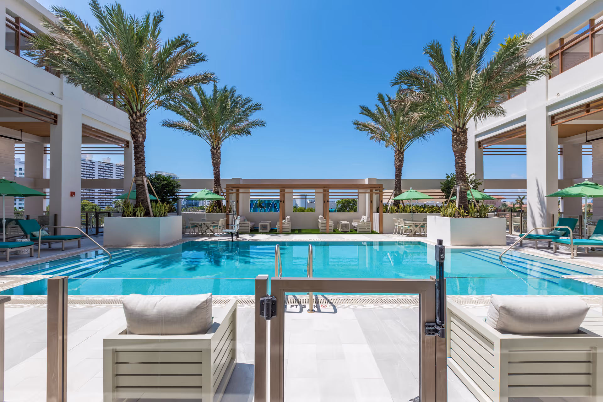 Outdoor swimming pool area with clear blue water, surrounded by palm trees and lounge chairs with green umbrellas under a bright blue sky.