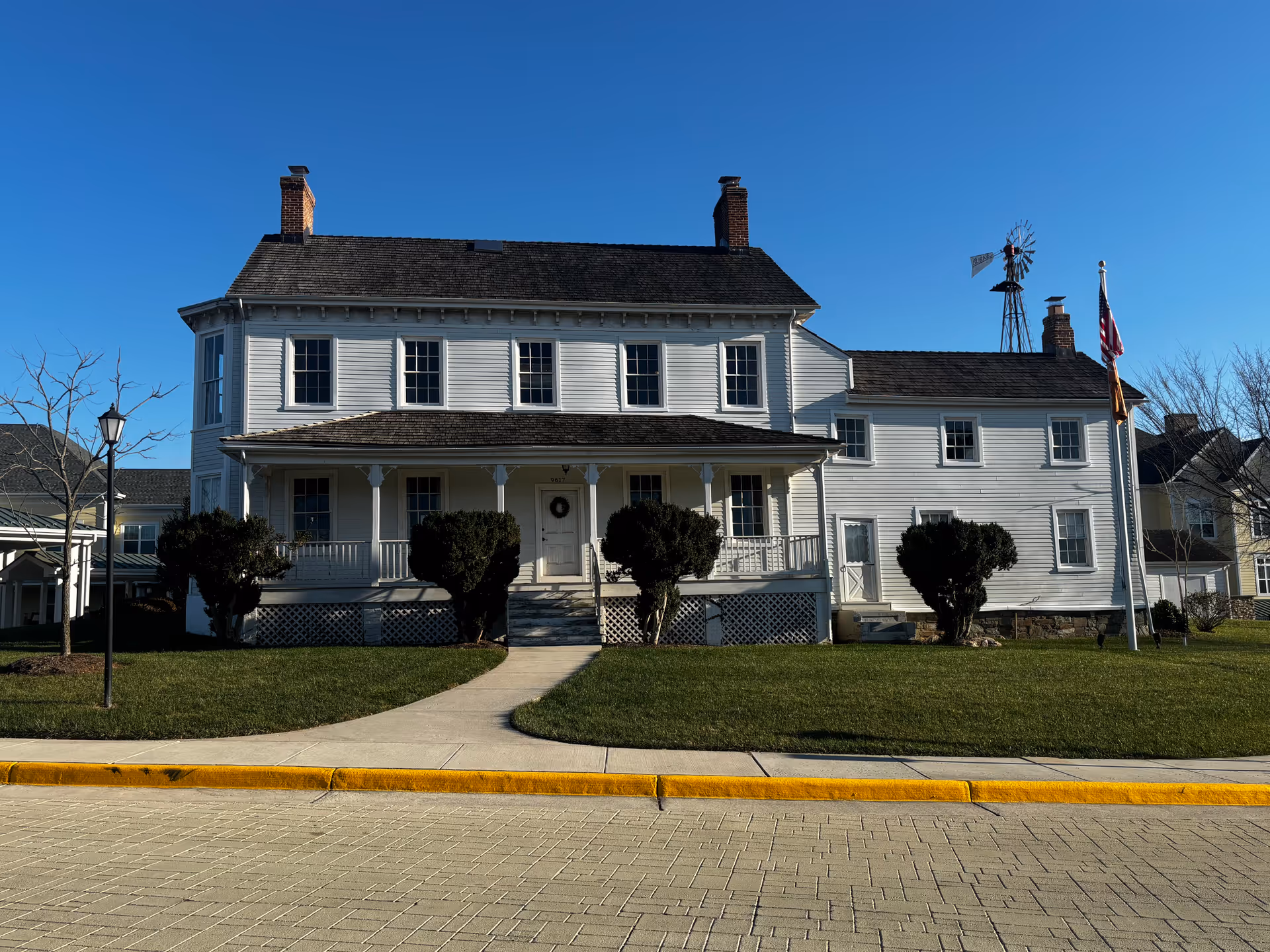 Front exterior view of a large white two-story house with a porch, multiple windows, and two chimneys. The house is surrounded by a well-maintained lawn with bushes and a flagpole with an American flag. A windmill structure is visible on the right side of the building under a clear blue sky.