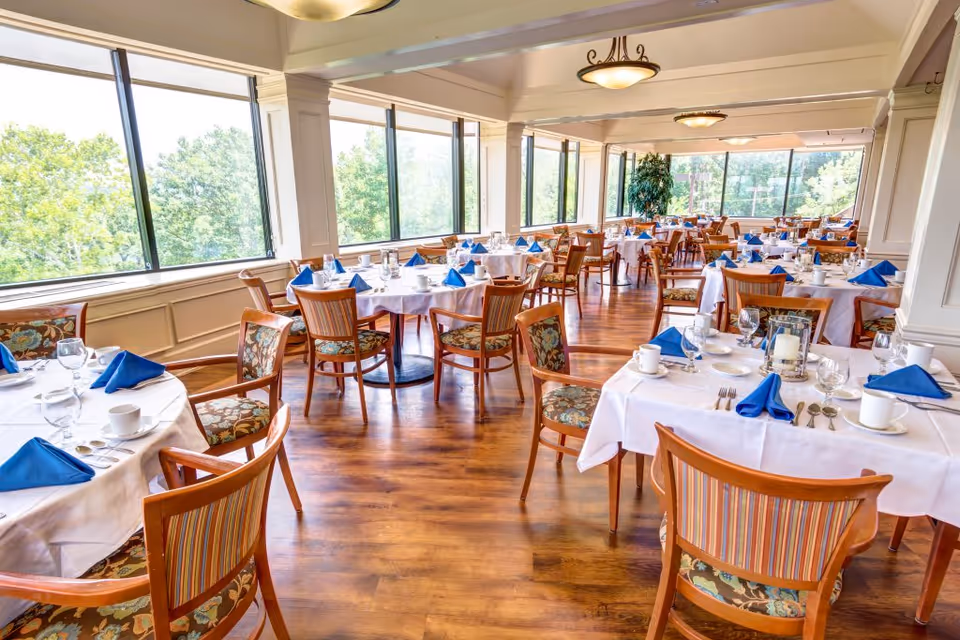 Bright dining room with large windows overlooking green trees. Multiple round tables are set with white tablecloths, blue folded napkins, glassware, cups, and silverware. Wooden chairs with patterned cushions surround the tables. The room has wooden flooring and ceiling lights.