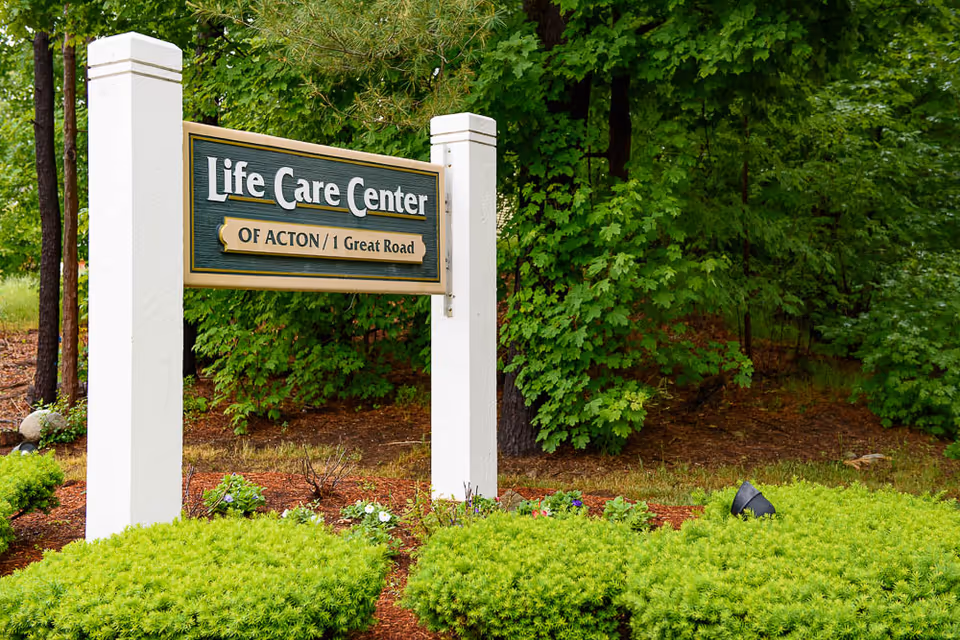 A wooden sign reading 'Life Care Center of Acton / 1 Great Road' mounted between white posts in a landscaped, tree-lined area.