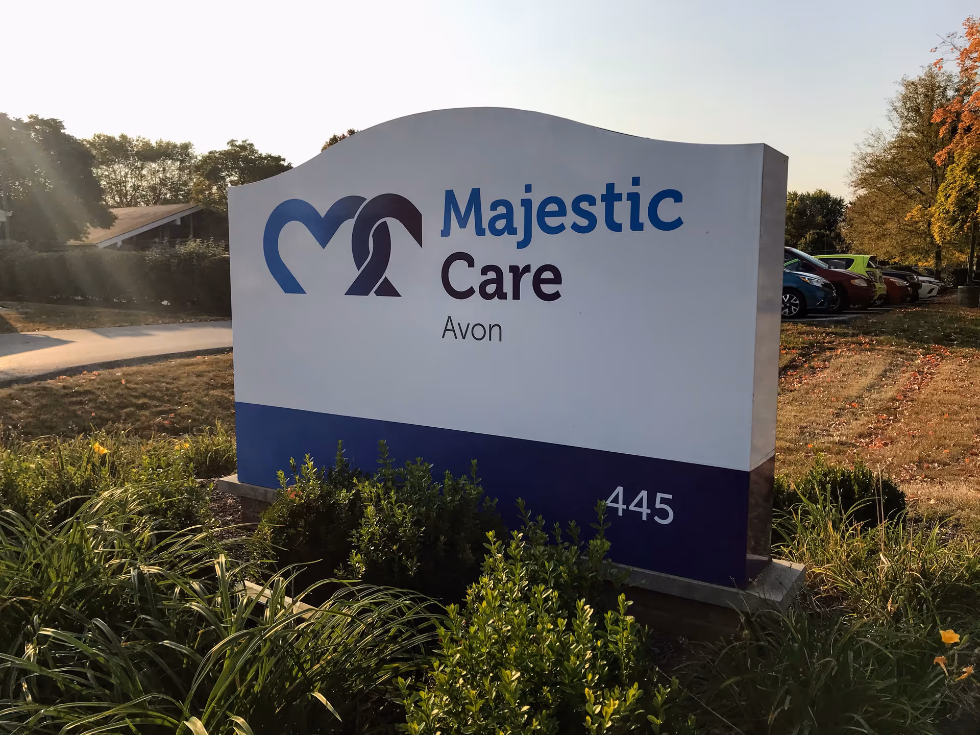 Outdoor sign for Majestic Care Avon with the address number 445, surrounded by greenery and a parking lot with cars in the background during daylight.