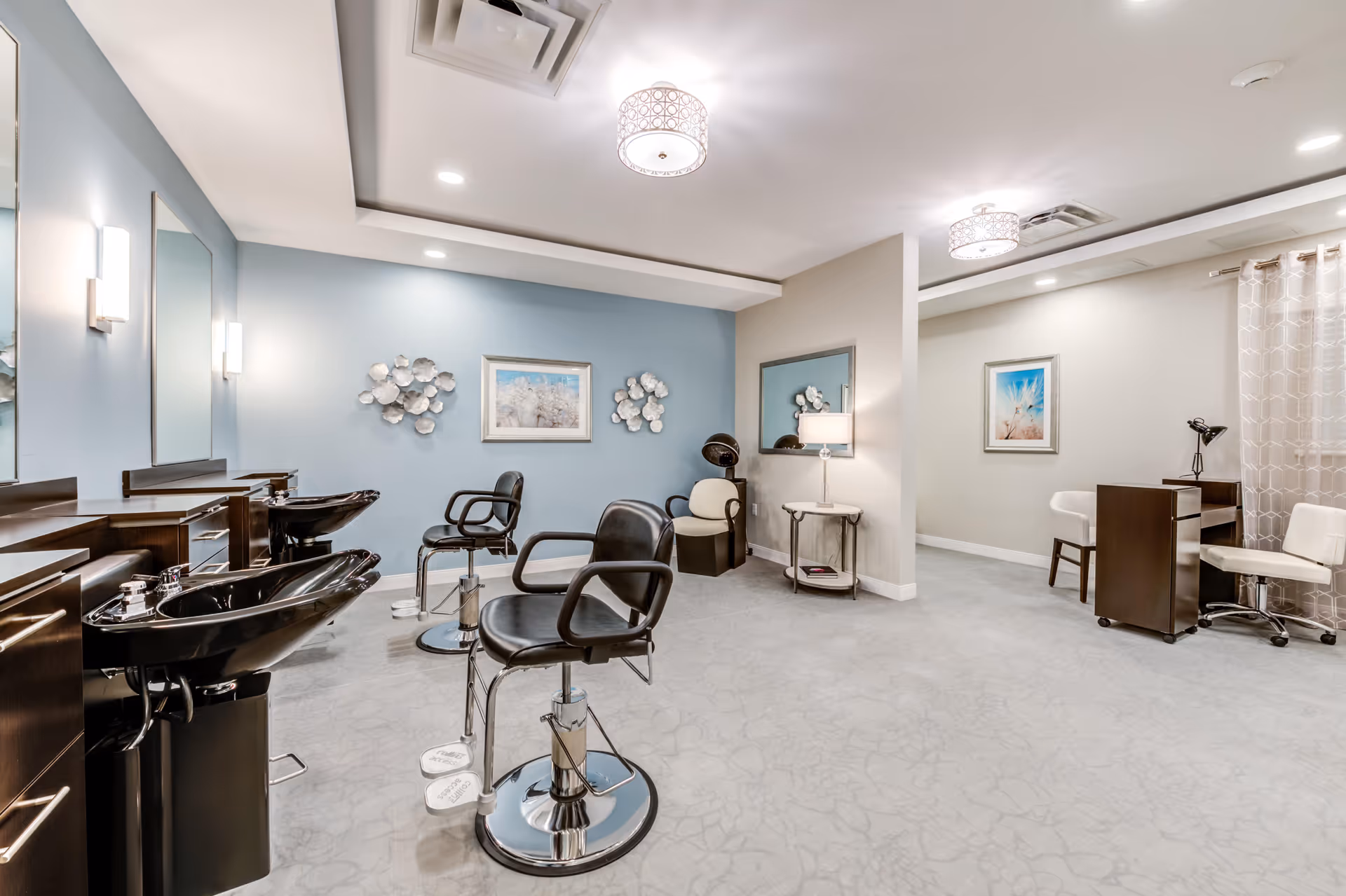 A modern salon area inside an assisted living facility with black salon chairs and sinks on the left, a hair dryer chair in the back, and a small desk with two white chairs on the right. The walls are painted light blue and beige, decorated with framed artwork and metal wall art. The room is well-lit with ceiling lights and wall sconces.