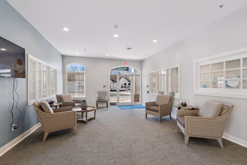 A bright and clean seating area inside a senior living facility with several cushioned wicker chairs arranged around small round tables. Large windows and glass doors allow natural light to fill the room. A flat-screen TV is mounted on the left wall, and the walls are painted in light gray and blue tones.