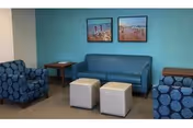 Small seating area with a blue sofa, two patterned armchairs, two white ottomans, side tables, and beach prints on a teal accent wall.