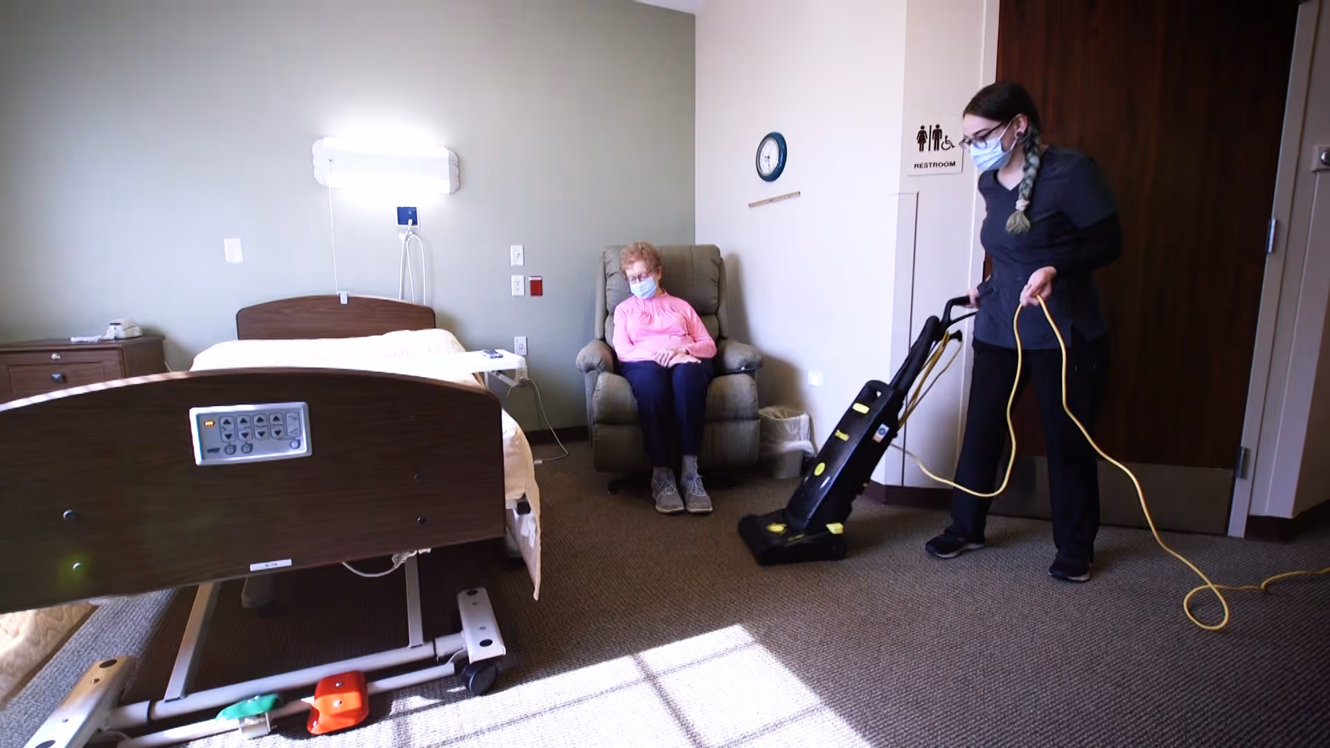 A senior woman wearing a mask sits in a recliner chair in a room with a hospital bed. A caregiver, also wearing a mask, is vacuuming the carpet. The room has a clock on the wall and a restroom sign near a door.