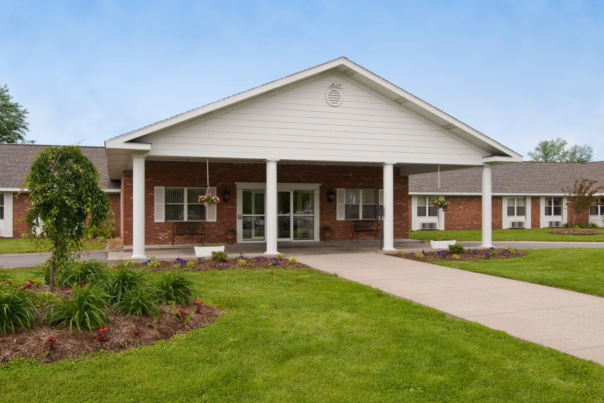 Front entrance of a single-story brick senior living building with white columns, a covered portico, walkway, and landscaped lawn.