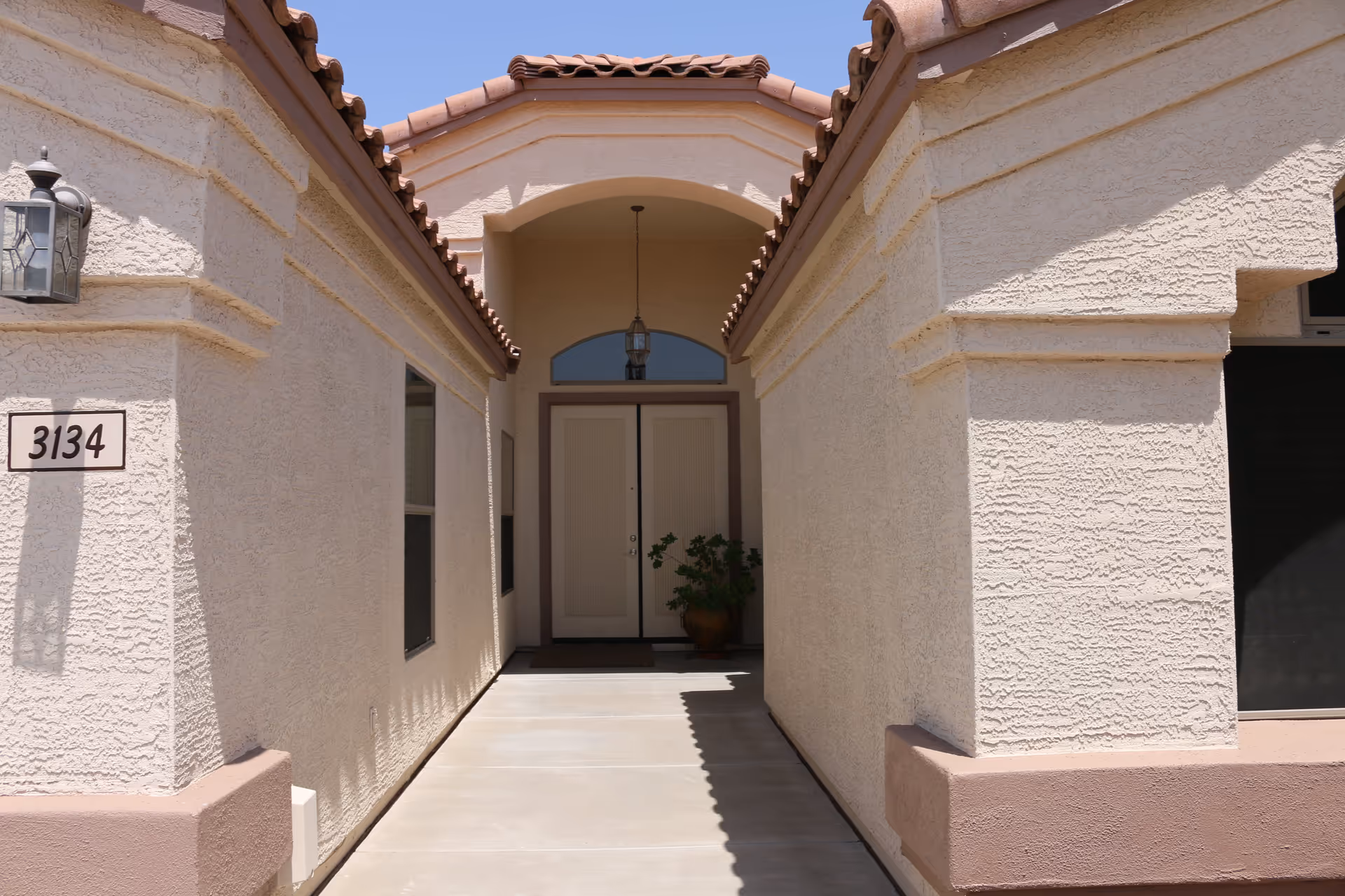 Entrance walkway leading to a double front door of a beige stucco building with a terracotta tiled roof. There is a potted plant near the door and a hanging lantern light fixture above the entrance. The building number 3134 is displayed on the left wall.