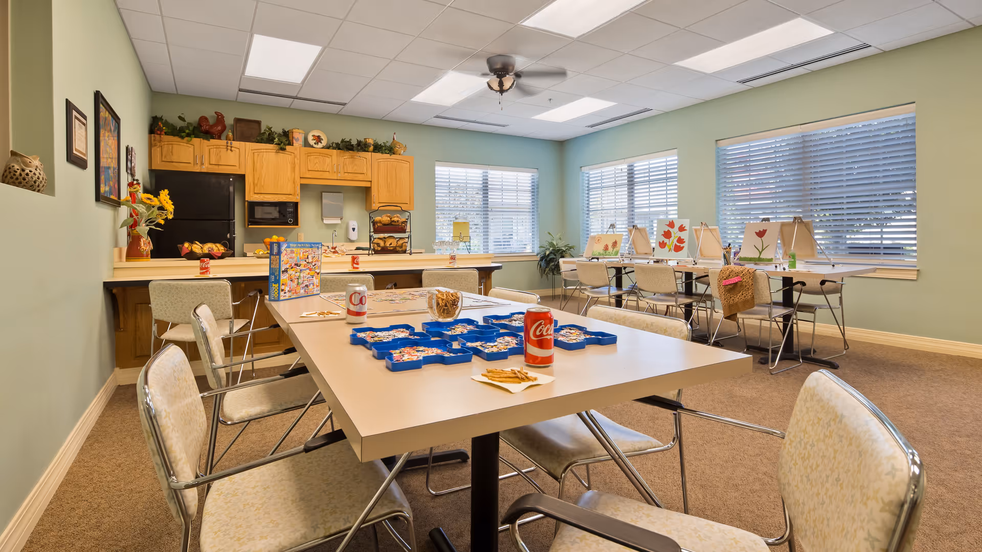 A well-lit activity room in an assisted living facility with tables and chairs arranged for group activities. One table has a puzzle in progress with puzzle pieces and soda cans on it. In the background, there are easels with paintings of flowers set up near large windows with blinds. The room has light green walls, a ceiling fan, and a kitchenette area with wooden cabinets and a black refrigerator.