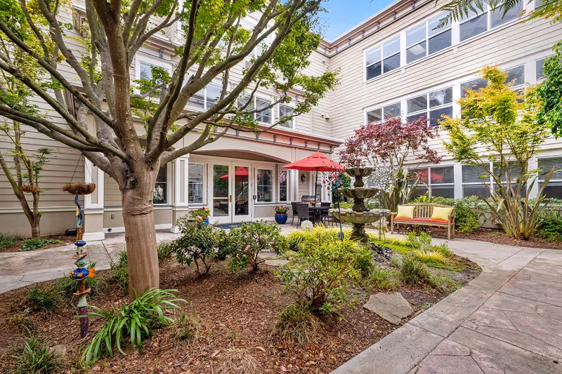 Outdoor courtyard area at Aegis Living San Francisco featuring a large tree, various shrubs and plants, a multi-tiered water fountain, a bench with yellow cushions, and a patio table with chairs under a red umbrella. The courtyard is surrounded by a three-story building with many windows.