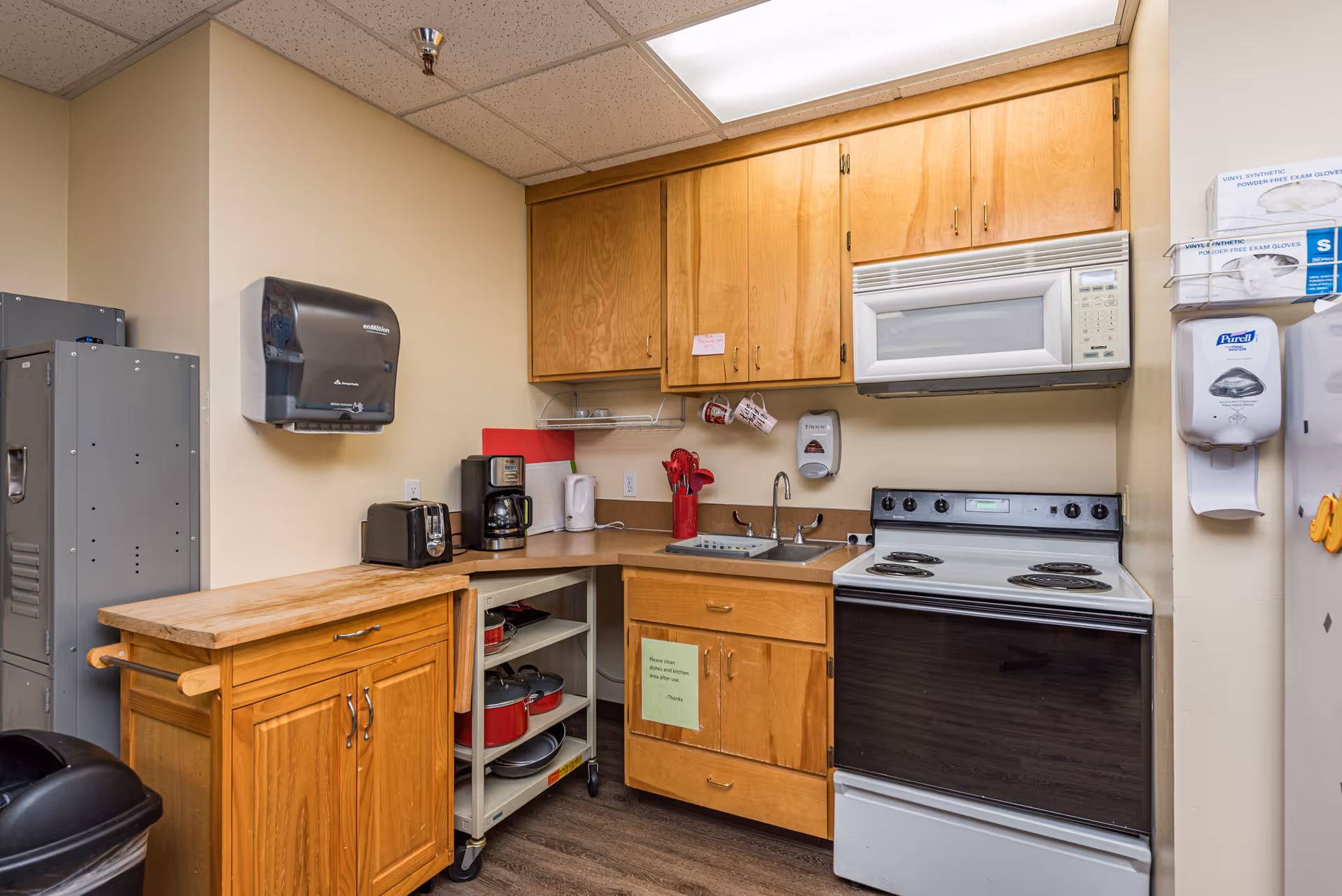 Small kitchen area with wooden cabinets, a white microwave above a white electric stove, a stainless steel sink, a coffee maker, toaster, electric kettle, and a wooden rolling cart with a butcher block top. There are paper towel and glove dispensers mounted on the wall, along with a hand sanitizer dispenser. The floor is wood, and the walls are painted beige.