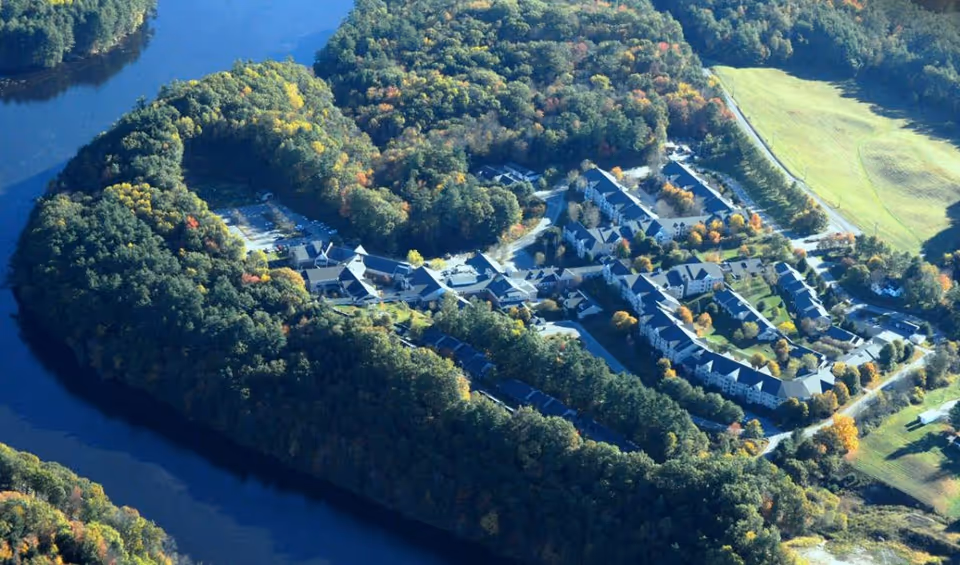 Aerial view of Kendal At Hanover facility surrounded by dense trees and bordered by a river, showing multiple buildings and parking areas within a wooded landscape.