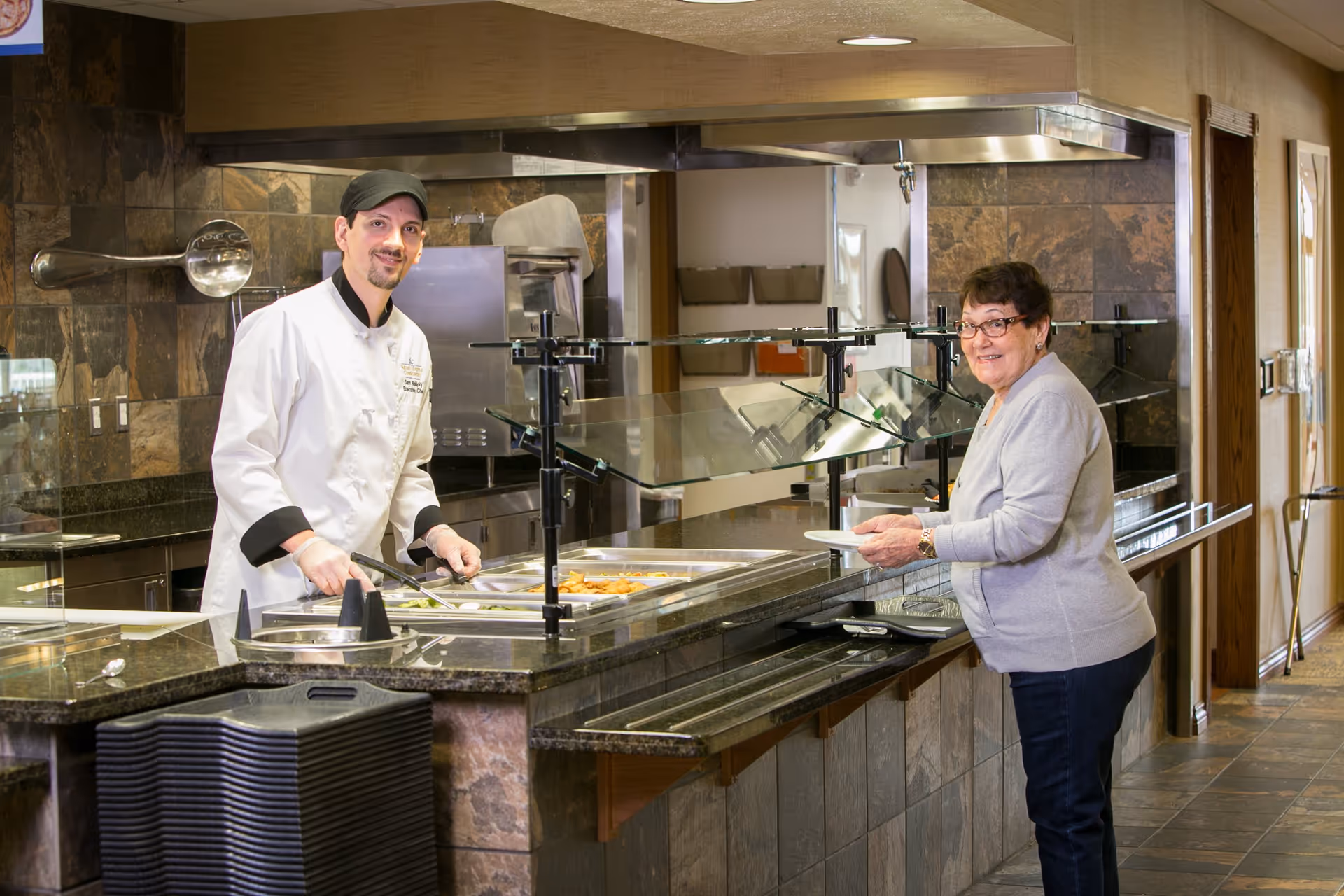 A chef wearing a white uniform and black cap is serving food from a buffet counter to an elderly woman holding a plate in a dining area with stone tile walls and a granite countertop.