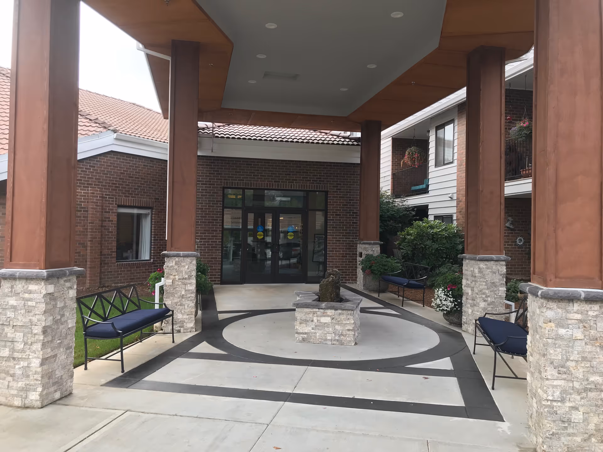 Covered entrance with large pillars, benches, a small central fountain, and glass double doors leading into a senior living building.
