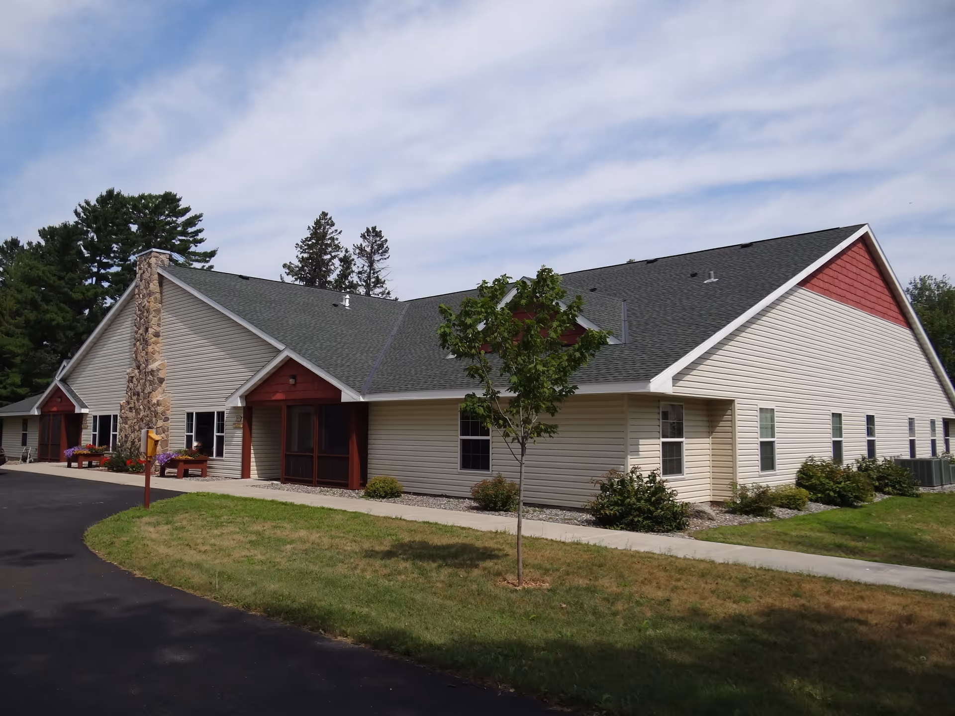 Exterior view of a single-story building with beige siding and a dark gray roof, featuring a stone chimney and red accents around the entrances. The building is surrounded by a lawn, a sidewalk, and some trees under a partly cloudy sky.