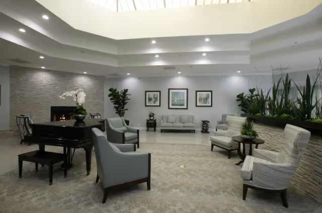 A spacious and well-lit living room area with a grand piano, several armchairs, a sofa, and a fireplace. The room features neutral tones, framed artwork on the wall, potted plants, and a skylight ceiling allowing natural light to enter.