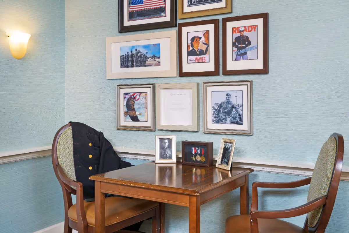 A small wooden table with two chairs in a corner of a room. One chair has a black jacket draped over it. On the table are three framed items: two photographs and a display case with medals. The wall behind the table is decorated with several framed pictures and posters, including images of military personnel, an American flag, and patriotic themes. The wall is painted light blue with a textured finish, and a wall sconce light is visible on the left side.