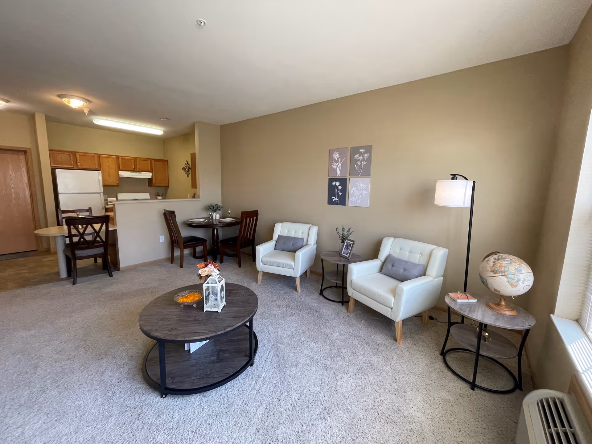 A cozy living room and dining area in an apartment with beige walls and carpeted floor. The living room has two white armchairs with gray cushions, a round wooden coffee table with a decorative lantern and flowers, and two side tables, one with a globe and lamp. The dining area features a small round table with two chairs and a kitchen with wooden cabinets, a white refrigerator, and a stove in the background.
