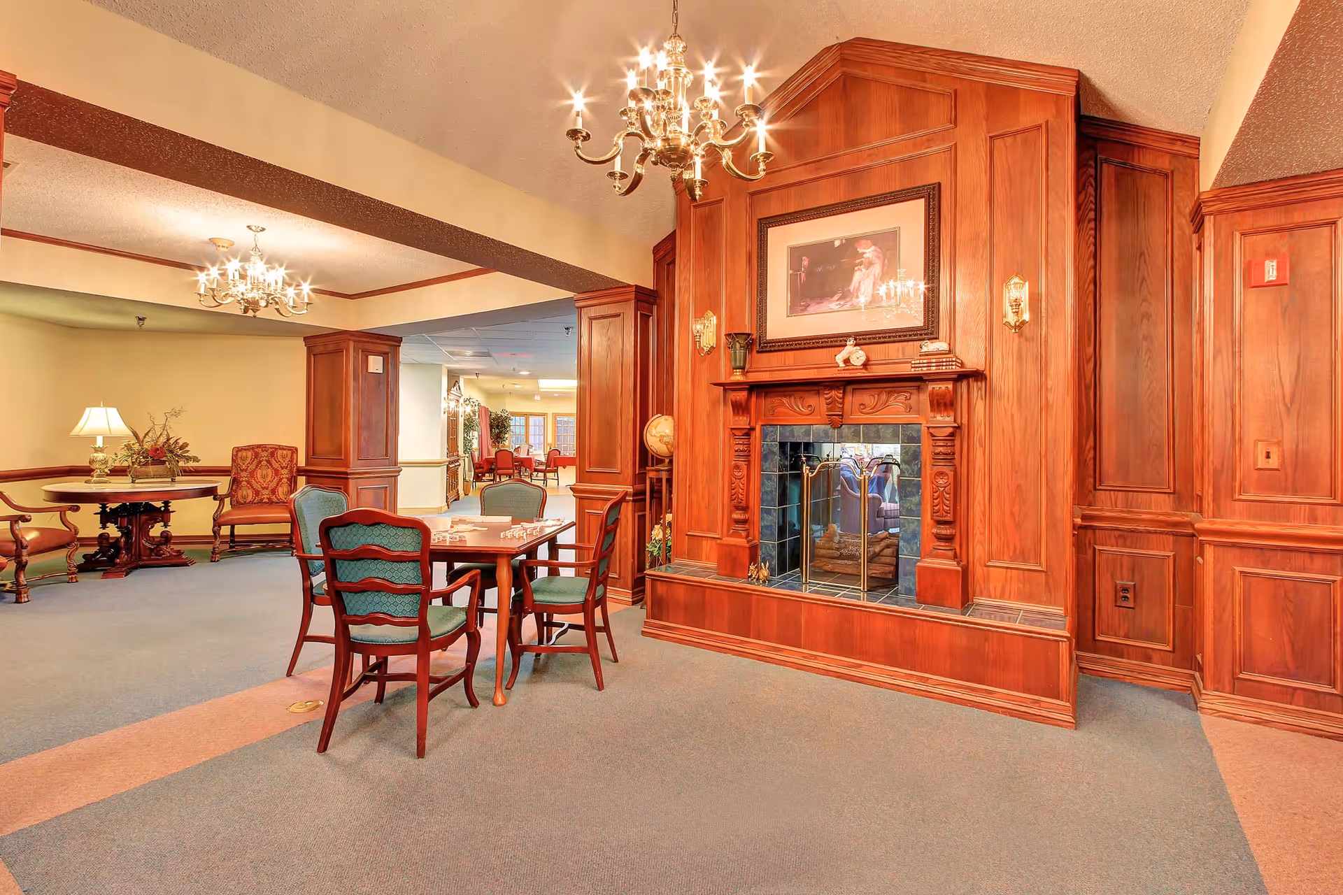 A cozy common area in a senior living facility featuring a wooden fireplace with a framed picture above it, a table with four chairs arranged around it, and additional seating and tables in the background. The room has warm lighting from chandeliers and lamps, with wood paneling on the walls and a carpeted floor.