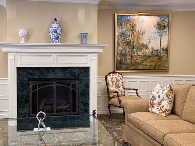 Cozy sitting area with a white mantel fireplace with green marble surround, upholstered chairs and sofa, glass coffee table, and a framed painting on the wall.