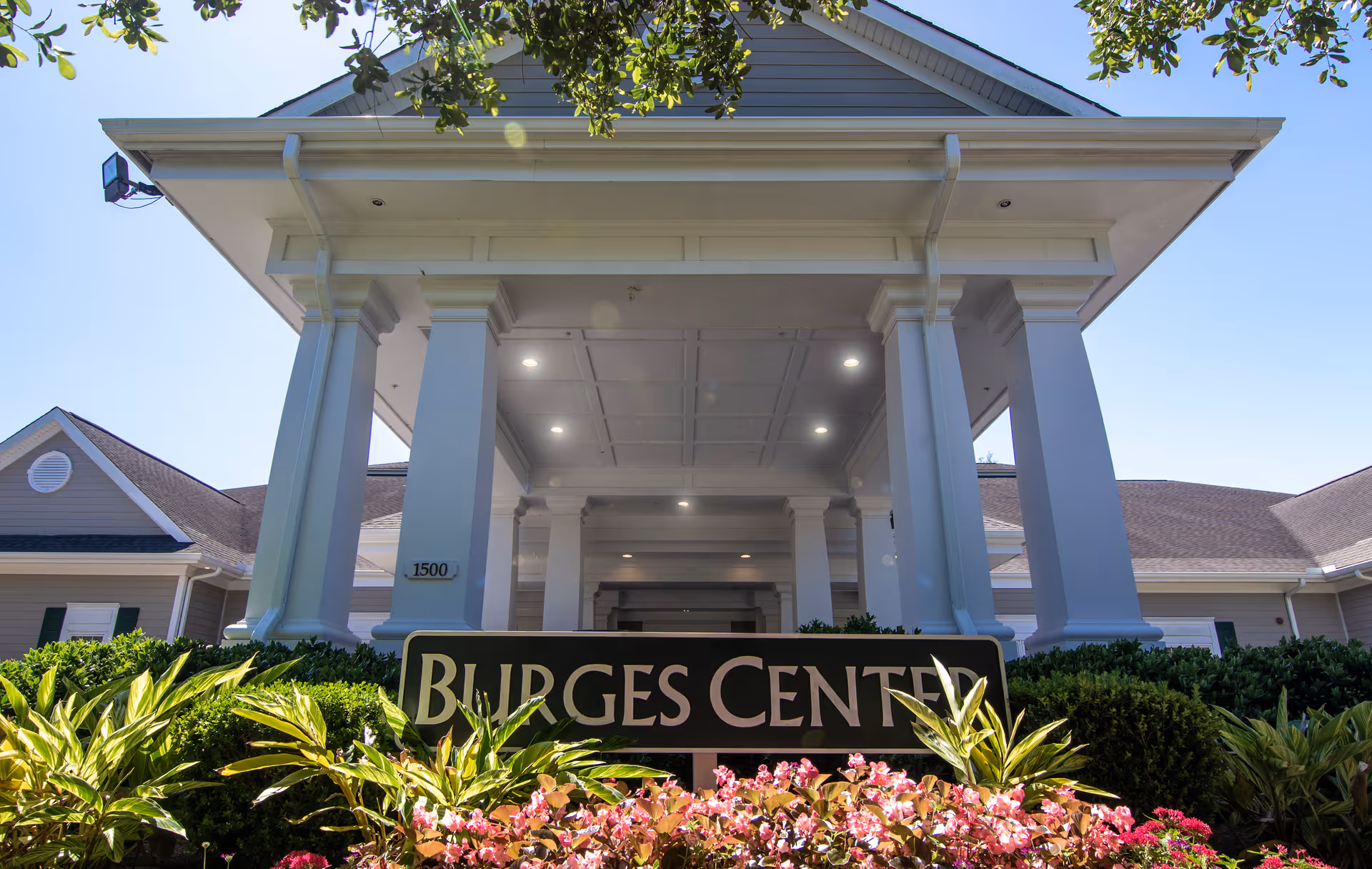 Front entrance of a building with large white columns, a covered portico and a sign reading 'Burges Center' surrounded by landscaping.