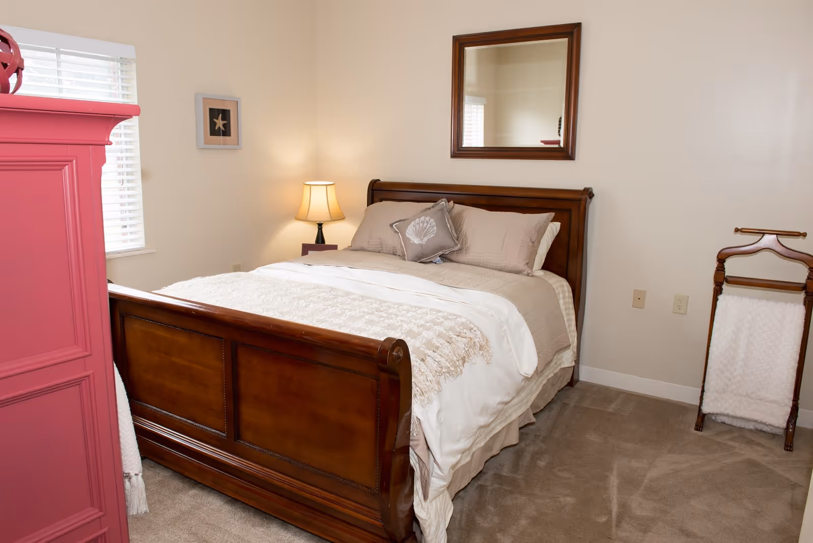 A cozy bedroom with a wooden bed frame, beige bedding, and a decorative pillow. There is a wooden nightstand with a lamp, a mirror hanging on the wall above the bed, a pink dresser on the left side, and a wooden towel rack with a white towel on the right side. The room has beige walls and carpeted flooring.