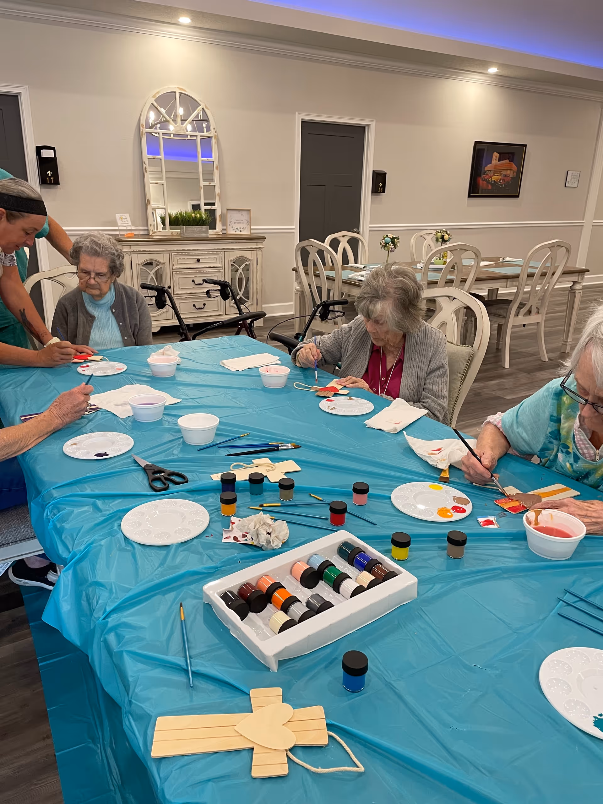 Several elderly women seated around a table covered with a blue plastic tablecloth, engaged in painting wooden craft items. Various paint pots, brushes, and palettes with paint are spread across the table. The room has light-colored walls, a mirror, and dining tables with chairs in the background.