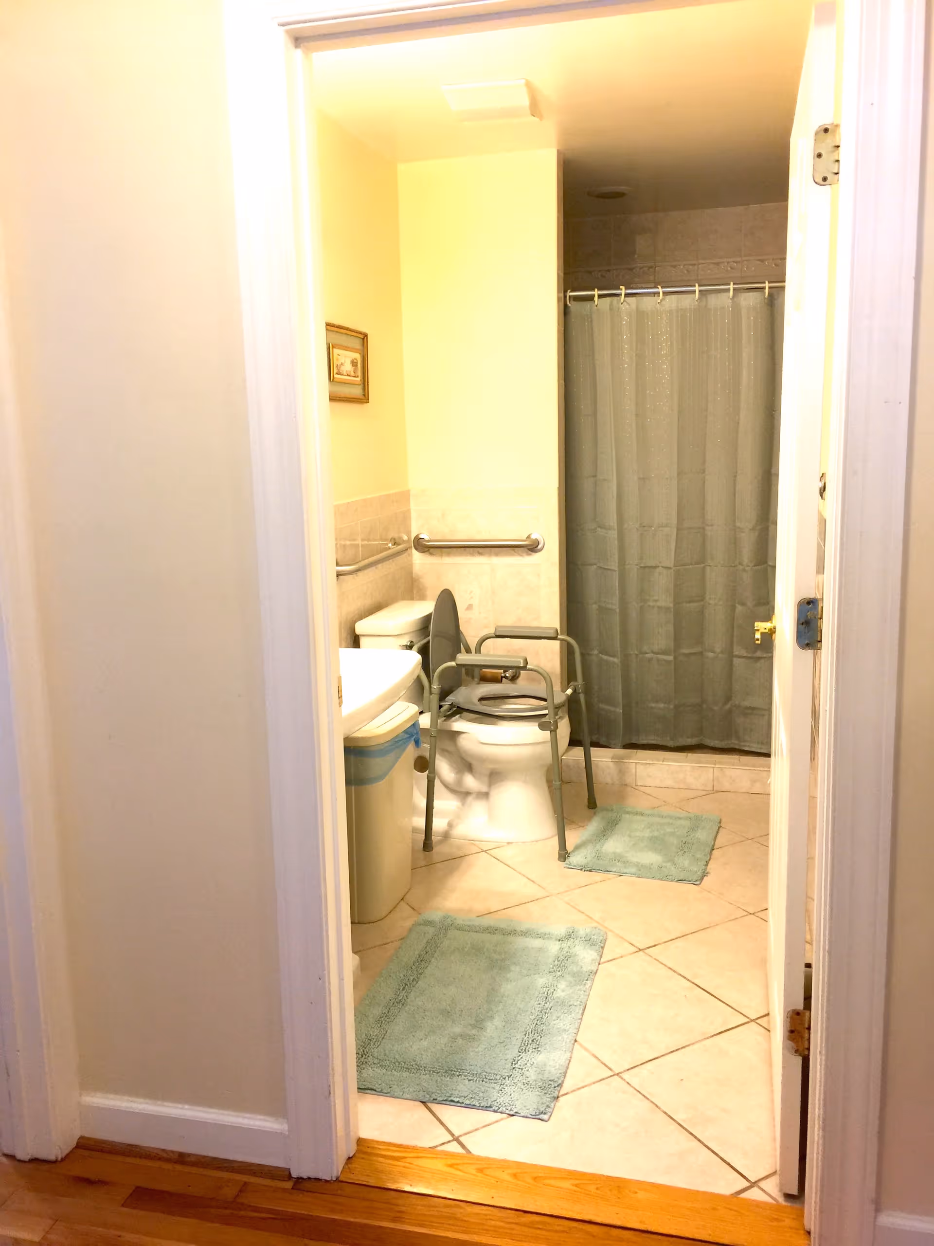 View into a bathroom with beige tiled floor and walls partially tiled. There is a toilet with a gray raised toilet seat and arm supports, a white sink with a trash bin underneath, a shower with a gray curtain, and two light blue bath mats on the floor. A grab bar is mounted on the wall next to the toilet.