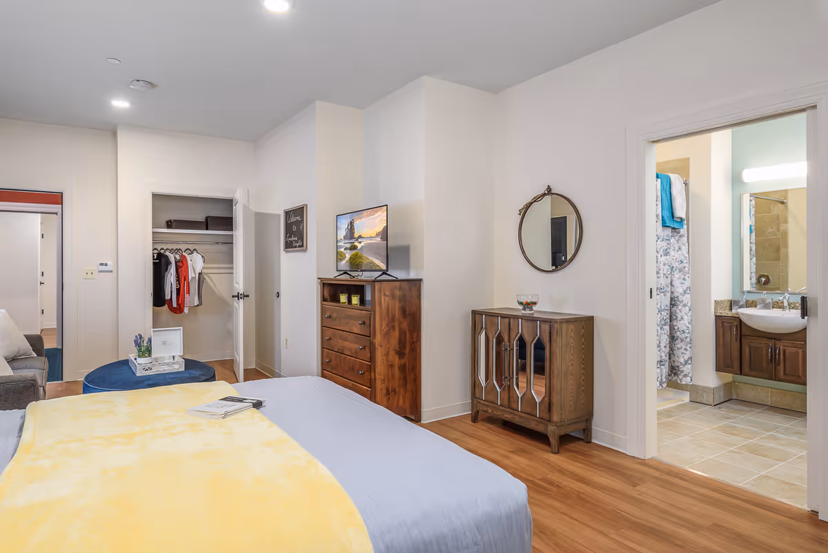 A bright bedroom with a bed covered in a yellow blanket and blue sheets, a wooden dresser with a TV on top, an open closet with hanging clothes, a small cabinet with a round mirror above it, and an open doorway leading to a bathroom with a sink and shower curtain visible.